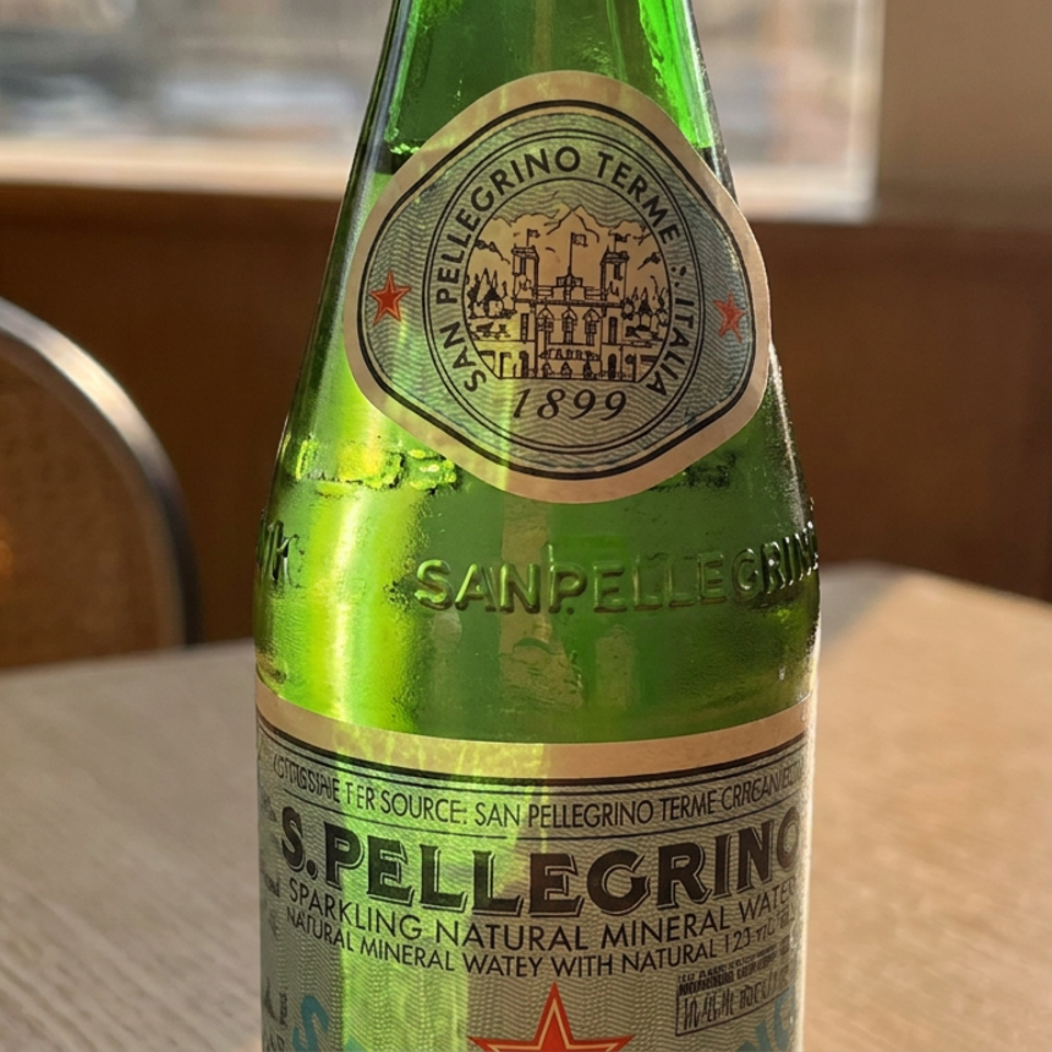 Green San Pellegrino sparkling mineral water bottle on a sunlit restaurant table beside a woven chair.
