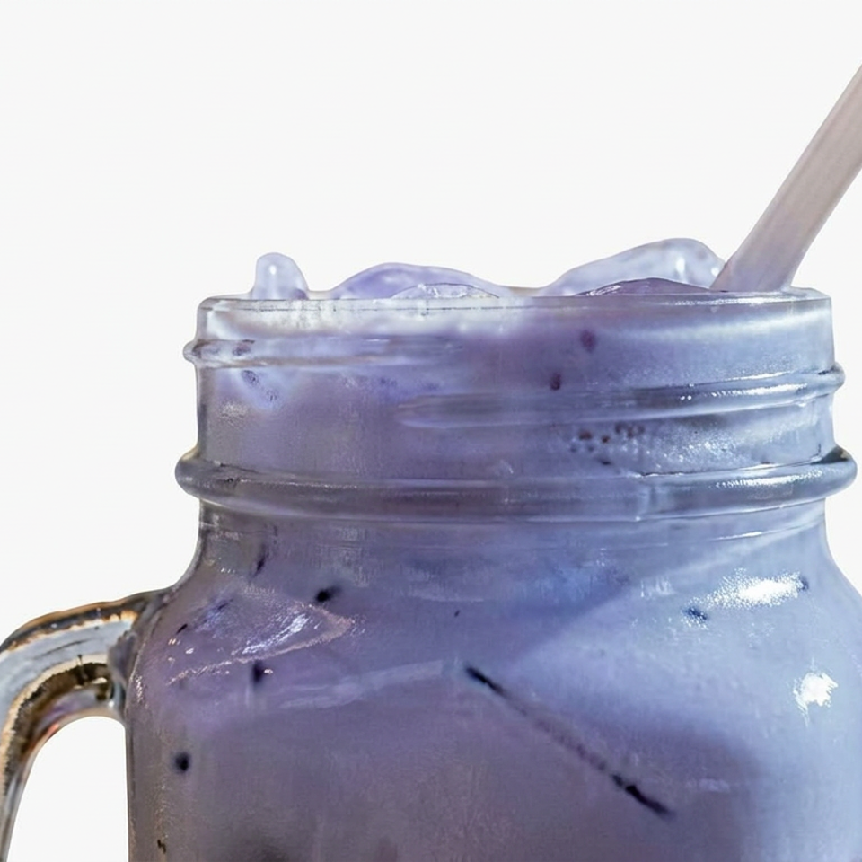 Close-up of a lavender-purple iced drink in a glass mason jar with a straw against a white background.