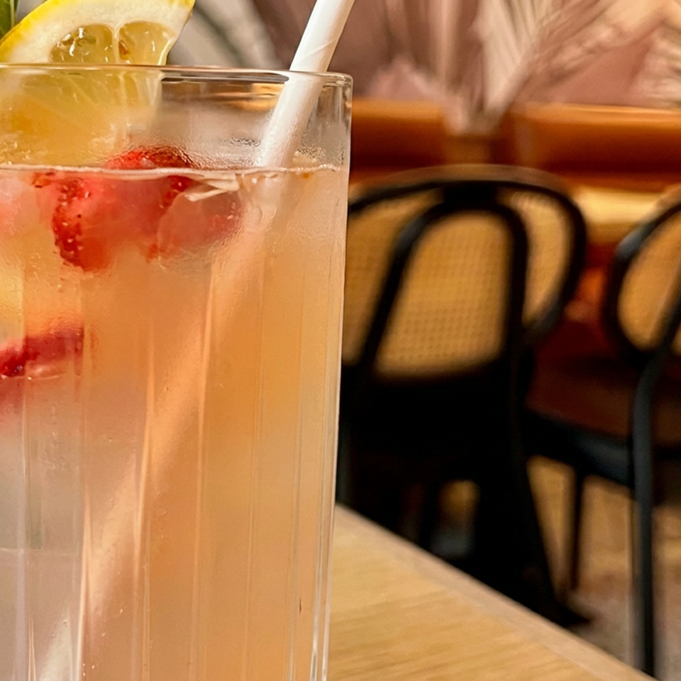 Tall glass of pink citrus drink garnished with lemon, mint, and berries, served with a straw in a warmly lit restaurant.